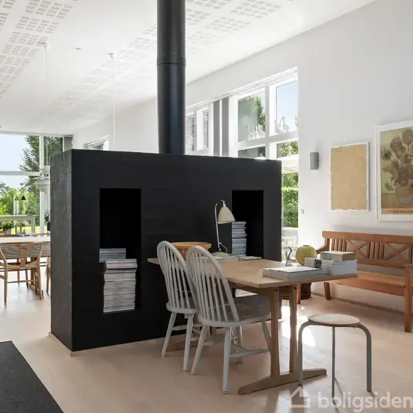 En spisebordsgruppe med stole og bord omkring en sort firkantet pejs, omgivet af lys moderne indretning med store vinduer og en bænk langs væggen.