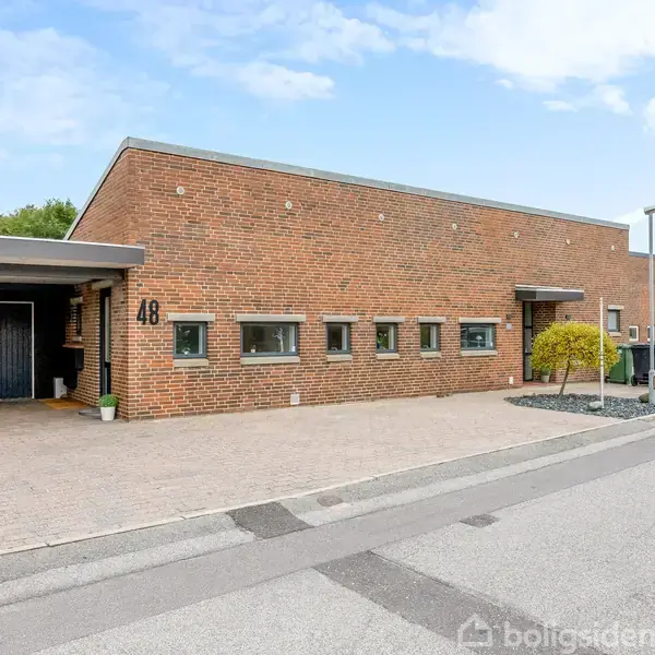 Rødt murstenshus med fladt tag, nummer 48, ligger ved en stille vej omgivet af andre lignende bygninger; indkørsel på venstre side fører til garage med sort dør.