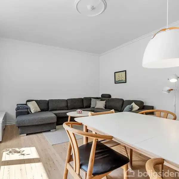En stue med en hjørnesofa og sofabord placeret ved en hvid væg. Et spisebord med stole står i forgrunden under en hængelampe. Væggene er hvide.