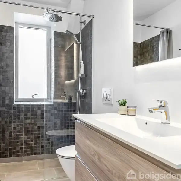 Et moderne badeværelse med glasbruseniche, vægmonteret toilet og træfrontet håndvask. Grå fliser på væggene, vindue for naturligt lysindfald, og håndklæder hænger ved siden af brusenichen.