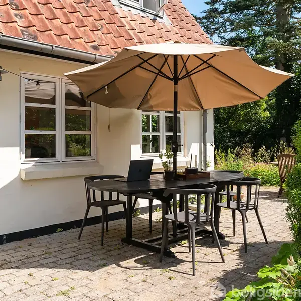 Terrassemøbler står under en parasol ved et bord på en stenbelagt terrasse foran et hus med rødt tegltag og hvide vinduer. Omkranset af grønne planter og træer.