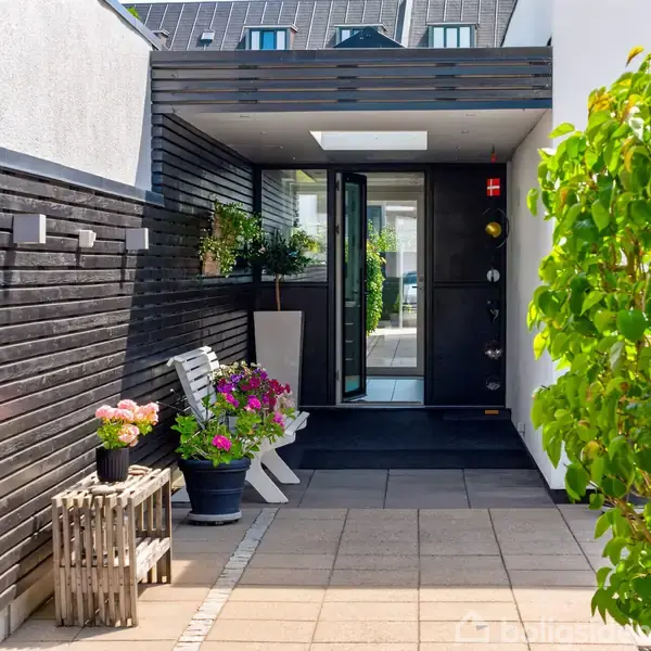 Indgangsområde med sort husfacade, hvor der står en bænk og en stol ved siden af blomstrende planter i potter. En grøn busk er placeret tæt på indgangen.