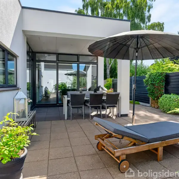 En solterrasse rummer en liggestol med parasol samt et spisebord med seks stole. Det er omgivet af grønne planter og flisebelagte overflader i en moderne have.