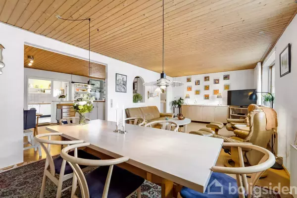 Langt spisebord med stole står centralt i en stue med træloft. Stuen har lædersofaer, planter og et tv. Åben adgang til køkken; lyse og varme trætoner dominerer rummet.