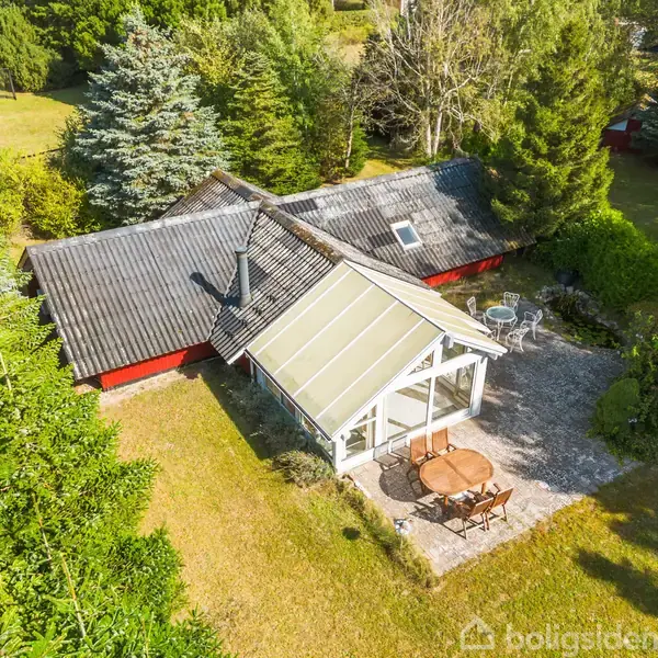 Et rødt sommerhus med en mørk tag ses fra oven, omgivet af tæt, grøn vegetation. Foran huset er der en terrasse med et rundt bord og stole.