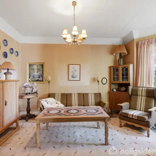 Stueindretning med en sofagruppe, træbord og to lænestole. Vægge dekoreret med tallerkener og malerier. Lampe hænger fra loftet; skænk står ved siden af en stor vindue.