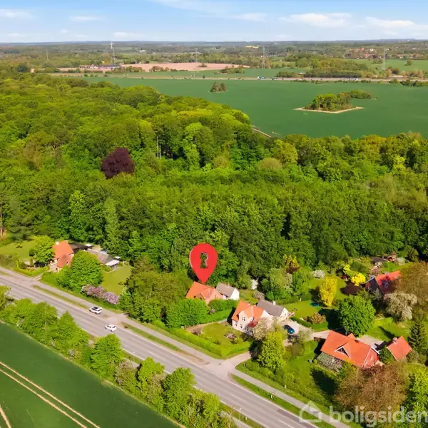 Et hus markeret med en rød nål ligger blandt grønne træer ved en vej. I baggrunden ses store marker og skove, der strækker sig mod horisonten.