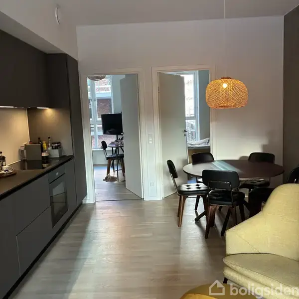 Et spisebord med stole står under en flettet lampeskærm. I baggrunden ses et moderne køkken til venstre, og en stue kan anes gennem åbne døre til højre.