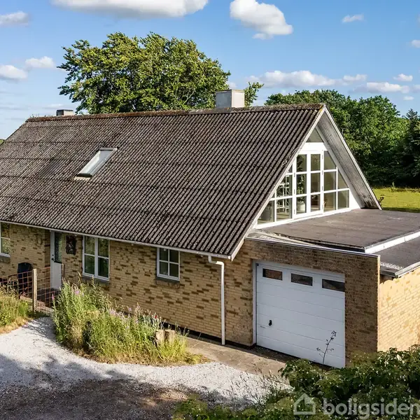 Et murstenshus med skråt tag har garage integreret. Det er omgivet af en grusvej og frodig grøn vegetation, mens skyer pryder den blå himmel i baggrunden.