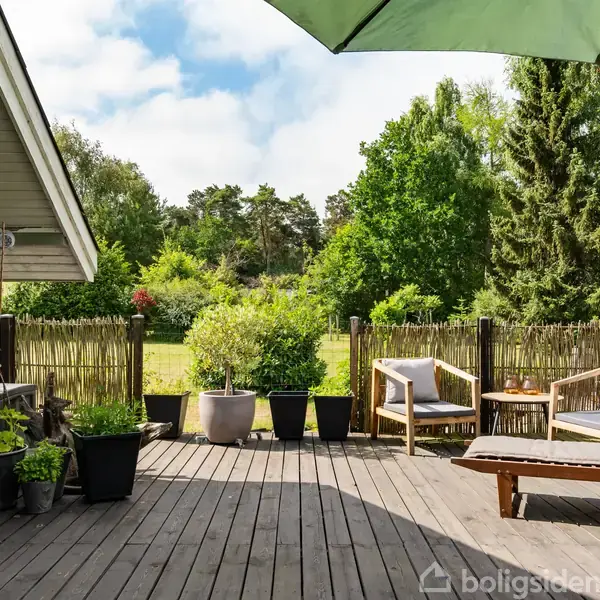 En træterrasse med solseng og møbler står under en stor grøn parasol, omgivet af blomsterpotter og grønne planter. Bagved ses en lukket, frodig have med træer.