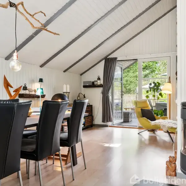 Spisebord med sorte stole står i en lys stue med skrå vægge. Ved siden af er en brændeovn. I baggrunden ses en åben dør til en grøn have.