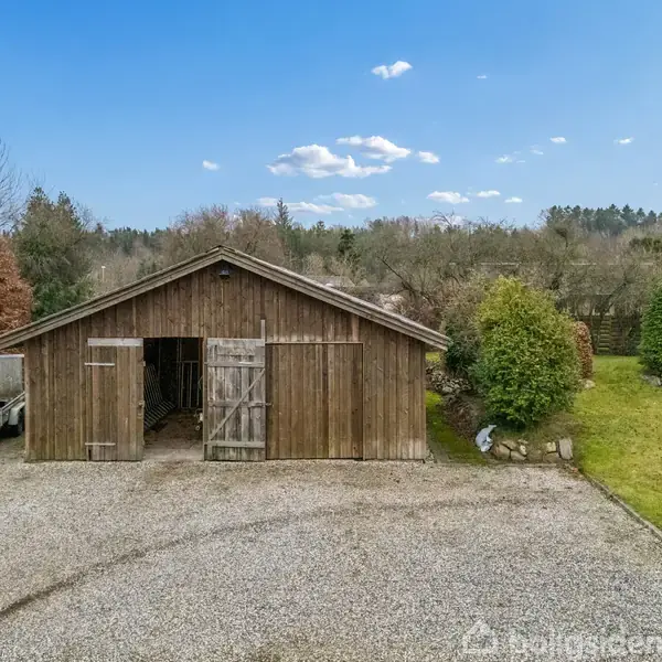 En stor, brun trælade står åben med døre, omkranset af grus og græsplæne i en grøn, frodig have med buske og træer.