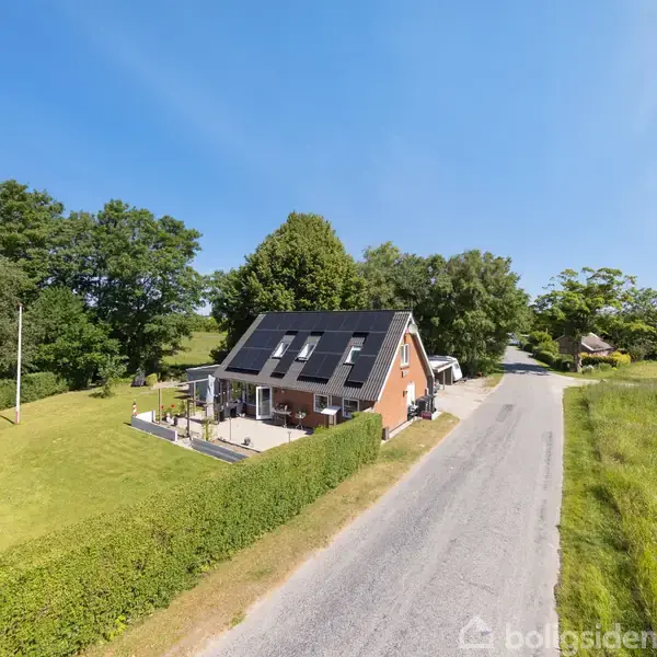Et rødt murstenshus med solpaneler står på en velplejet græsplæne. Huset er omgivet af en hæk og en landevej, med skov i baggrunden og marker til højre.