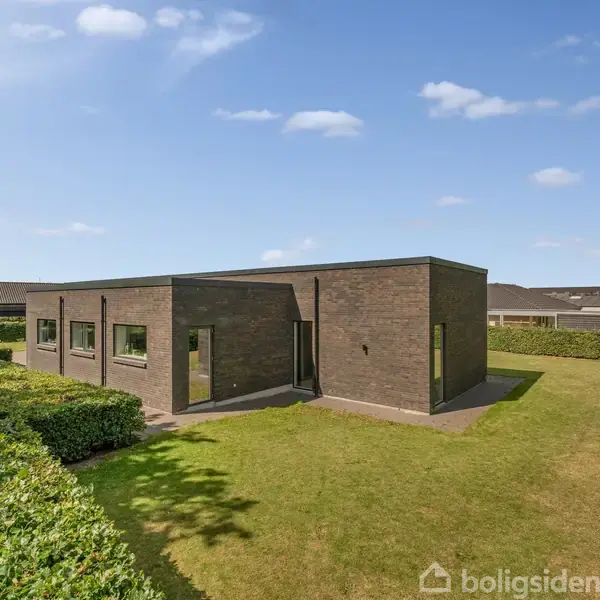 En enkel, rektangulær, mørk murstensbygning står midt i en velplejet græsplæne. Bygningen har flere vinduer, og en sti omgivet af hække fører til huset. Himlen er blå med få skyer.