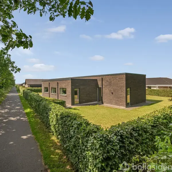 En moderne, enkelt-etage bygning står stille omringet af en velholdt hæk og græsplæne. Det er placeret ved siden af en asfalteret sti omgivet af træer under en klar blå himmel.