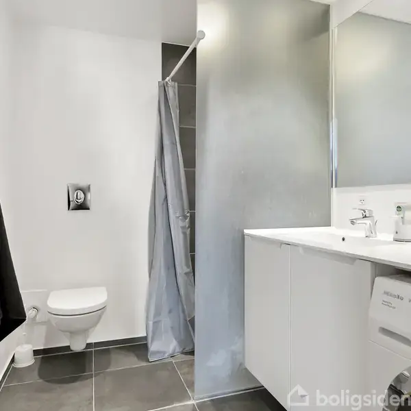Badeværelse indeholder toilet, bruser med forhæng, vask med spejl og vaskemaskine. Flisebelagt gulv og hvidmalede vægge skaber et rent og moderne udtryk i rummet.