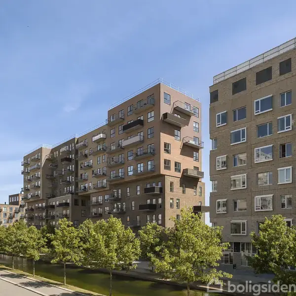 Flere moderne lejlighedskomplekser står langs en træbeplantet gade under en klar blå himmel. Bygningerne har forskellig arkitektur med balkonudformninger og store vinduespartier.