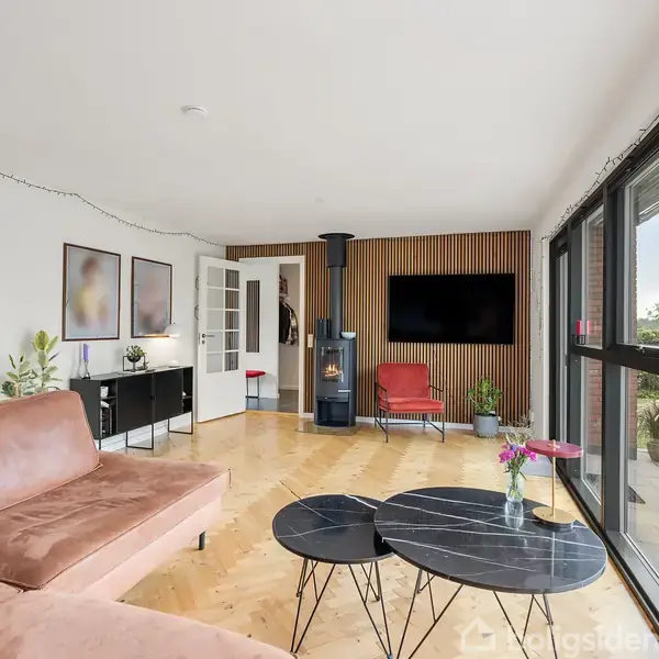 Stue med brun sofa, runde sorte borde og brændeovn ved væggen. Vinduer med udsigt til have. Væg dekoreret med billeder og sort skænk. Blomster i vase på bordet.