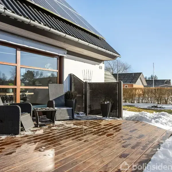 Terrassesæt med stole og bord står på en solrig, træbelagt terrasse ved et hus med store vinduer. Sne dækker græsplænen i baggrunden. Husnummer 40 vises på facaden.