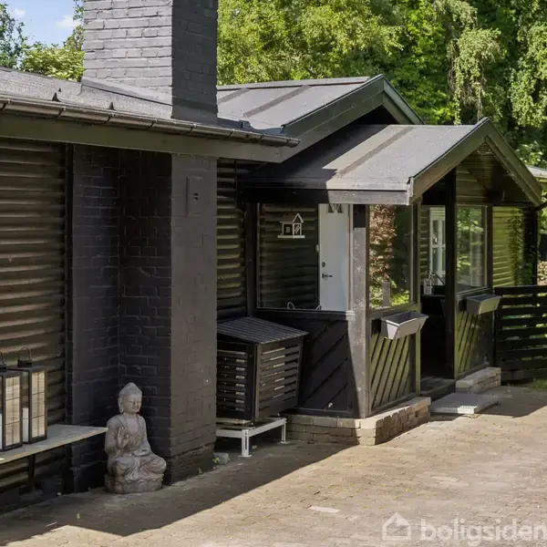 Et sommerhus med mørkt træbeklædning står stille i en skovklædt have. På en bænk ved siden af huset sidder en Buddhalignende statue. Sollys filtrerer gennem de omkringliggende træer.