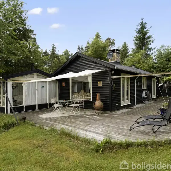 Sort, træbeklædt sommerhus med store vinduer, står på en træterrasse med havemøbler og liggestol. Det er omgivet af grønne træer og buske på en græsplæne.