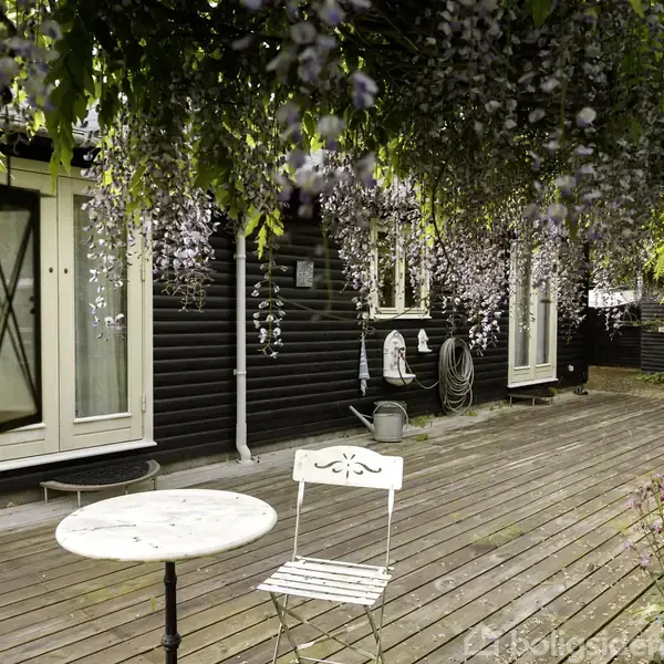 Rundt havebord og stol står på en træterrasse omgivet af hængende lilla blomster. En lanterne hænger ned fra loftet. Baggrunden viser en sortmalet husfacade med hvide døre.