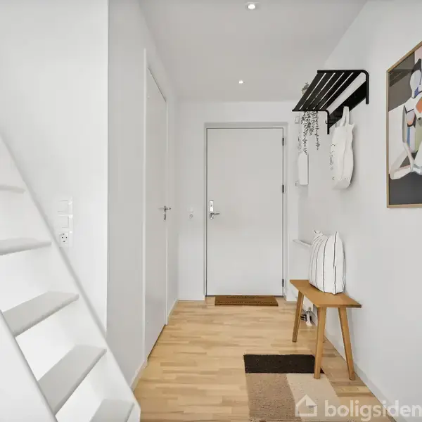 En lys entré med en hvid trappe til venstre. En bænk med puder står til højre under en billedramme. En dør for enden omgivet af minimalistisk indretning.
