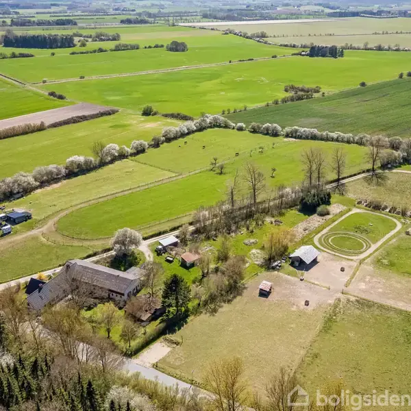 Luftfoto af en gård med store bygninger, der står stille i et frodigt, landligt område omgivet af marker og træer. Flere marker og stier kan ses i det åbne landskab.