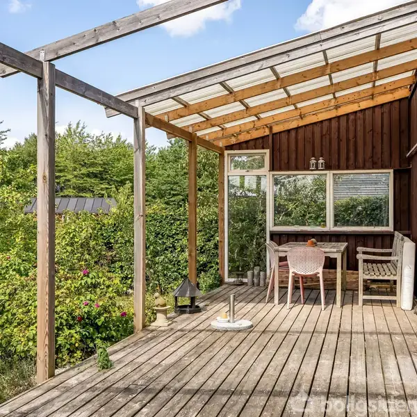 Terrasse med trædæk, overdækket af en pergola. Et bord og to stole står nær husmuren, omkranset af frodige træer med røde æbler. Sollys og grønne buske omgiver stedet.