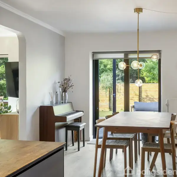 Spisebord omgivet af stole står i spisestue med lyse vægge. Et klaver ved siden af bordet. Store vinduer åbner mod en terrasse med grønne planter.