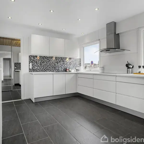 Moderne køkken med hvide skabe og sort flisegulv. Mosaikfliser på væggen bagved vasken. Omgivet af to store vinduer, der giver lysindfald. Ingen tekst påvist.