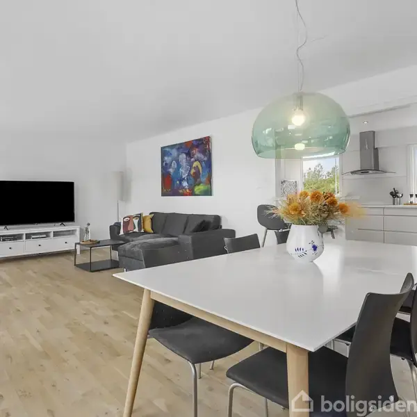 En moderne stue med spisebord, blomstervase og sorte stole. Sofa og fladskærms-TV er i baggrunden. Farverig kunst hænger på væggen, og køkkenområdet ses til højre.