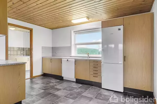 Et køkken med træloft og grå flisegulv. Hvide skabe med træpaneler omgiver en opvaskemaskine og et hvidt køleskab. Et stort vindue slipper naturligt lys ind.