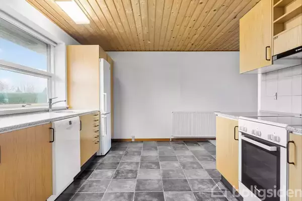 Et køkken med lyse træskabe står tomt. Køleskab, ovn og opvaskemaskine er til stede. Kvadratiske, mørke gulvfliser dækker gulvet. Et stort vindue giver udsigt til et udendørs område.