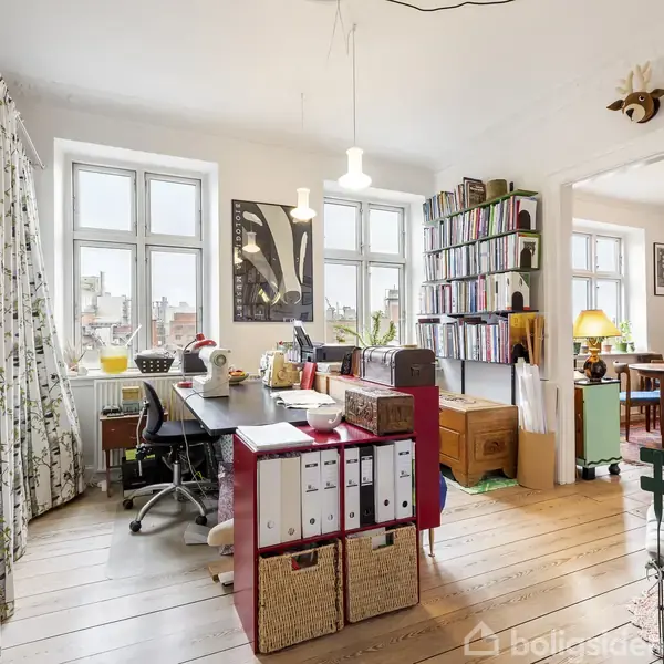 Et hjemmekontor med et stort skrivebord fyldt med kontorartikler og papirer. Gardiner hænger ved vinduer, og reoler fyldt med bøger og mapper står ved væggen. Ved siden af er et spiseområde.