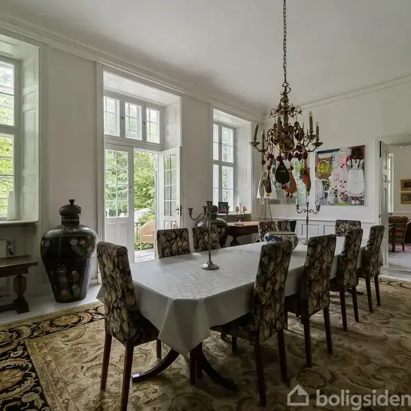 Spisebord dækket med dug og stearinlys, omgivet af polstrede stole. En lysekrone hænger over bordet. Store vinduer lader naturligt lys strømme ind i det elegante rum.