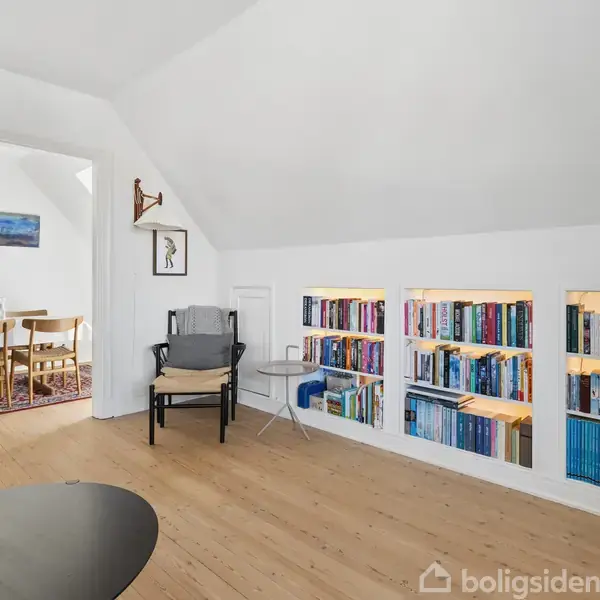 Hylde med bøger er indbygget i væggen, belyst med indbygget lys. I rummet står en grå stol med sofabord ved siden af. Åben adgang til spisestue med træstole.
