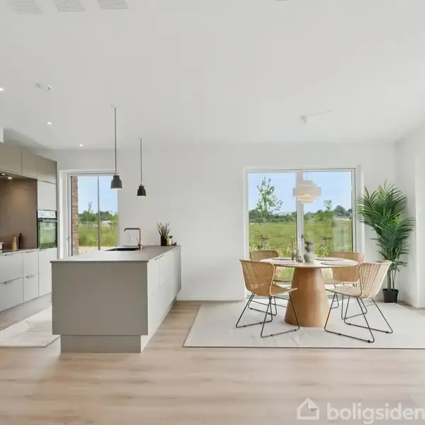 Køkken med gråt køkkenskab og ø, til venstre. Spiseområde med rundt træbord og fire stole, midt i et lyst rum med udsigt til natur gennem store vinduer.