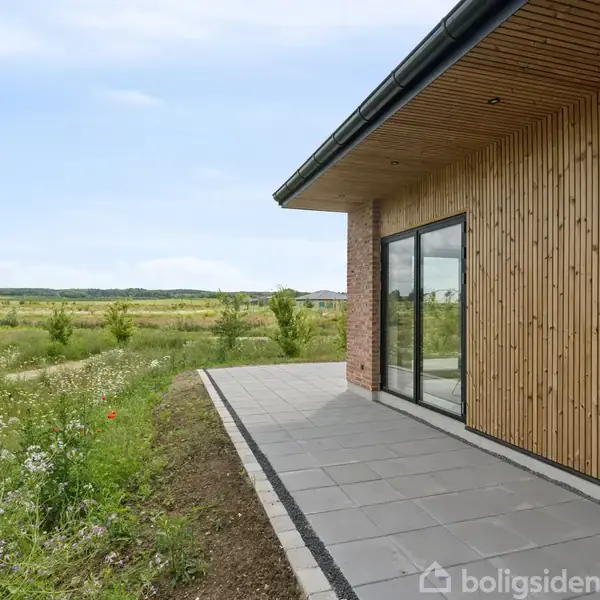 Hus med træbeklædning står stille, omgivet af en flisebelagt terrasse. Det grønne område strækker sig i baggrunden under en klar blå himmel.