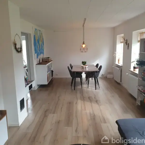 Spisebord med stole står på trægulv under en hængende lampe. Omgivet af hvide vægge med kunst, reoler med bøger og vinduer, der lader naturligt lys komme ind.