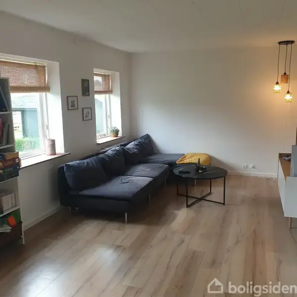 En stor, blå sofa står i et lyst stuehjørne ved siden af et rundt sofabord. Hylder med bøger og spil står ved vinduerne, mens lamper hænger fra loftet.