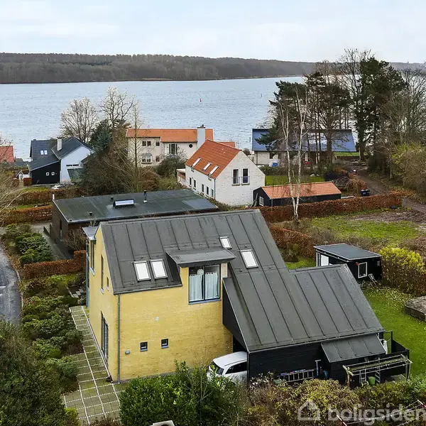 Gult enfamiliehus med sort tag står stille i forstadsområde ved vandet. Omkringliggende huse og træer rammer nabolaget med udsigt over en bred sø eller fjord.
