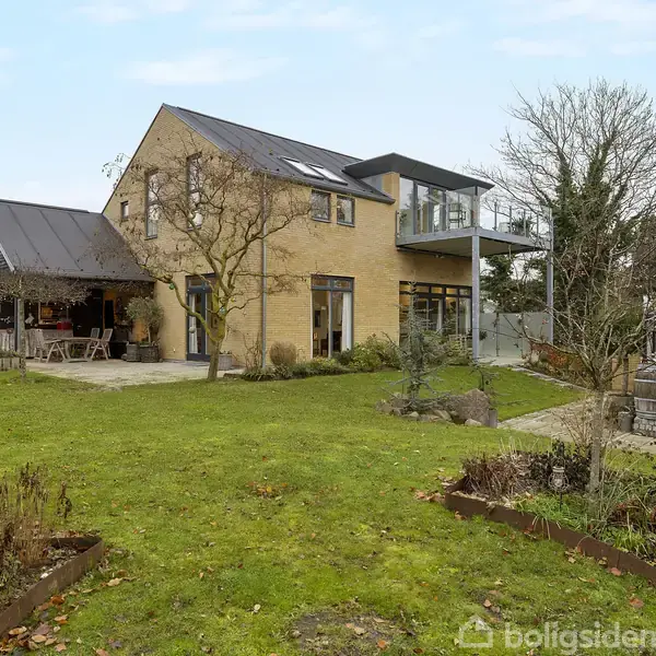 To-etagers murstenshus med store vinduer og terrasse, omgivet af en stor have med træer og en pavillon. Et udendørs spiseområde er placeret under et overdækket område ved huset.