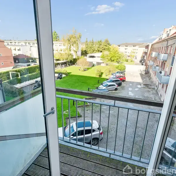 Glasdør åbner ud til en lille balkon med udsigt over en parkeringsplads og græsareal. Flere biler er parkeret, og bygninger omkranser området under en blå himmel.