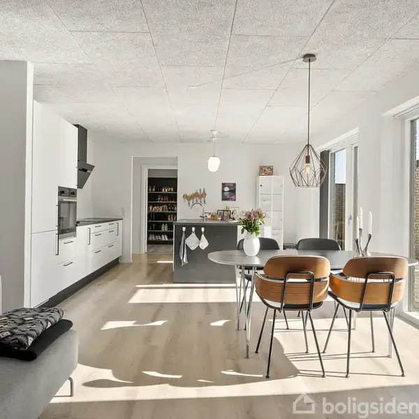 Køkken-alrum med spisebord og fire stole ved store vinduer, grå sofa i forgrunden. Køkkenet har hvide skabe og grå bordplade. Udenfor ses terrasse. Vægtekst: "Livet tager..." (resten udeladt).