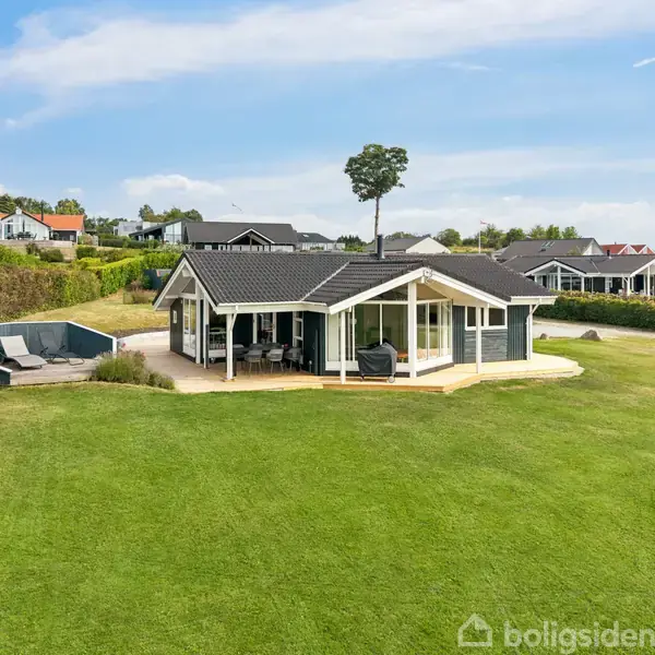 Et moderne sommerhus med store vinduer står på en velplejet græsplæne. En terrasse med havemøbler ligger ved siden af huset. I baggrunden ses flere sommerhuskonstruktioner i et naturskønt område.
