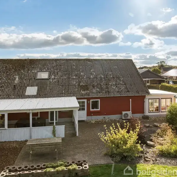Rødt hus med halvanden tag, forbundet til en carport. Omkring huset er der en grus- og haveområde med buske og små træer under en klar, delvist skyet himmel.