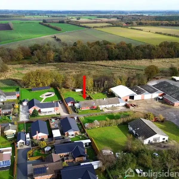 Et hus markeret med en rød pil står blandt flere bygninger. Omgivet af marker og træer i et landligt område med spredte bebyggelser og nogle landbrugsejendomme.