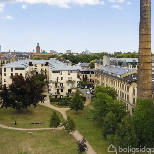 Udendørs terrasse med sort bord og stole, omgivet af hæk og planter. En altan hænger ovenover. Skiltet ved vinduet siger "til salg" med logoet af Boliga.dk.