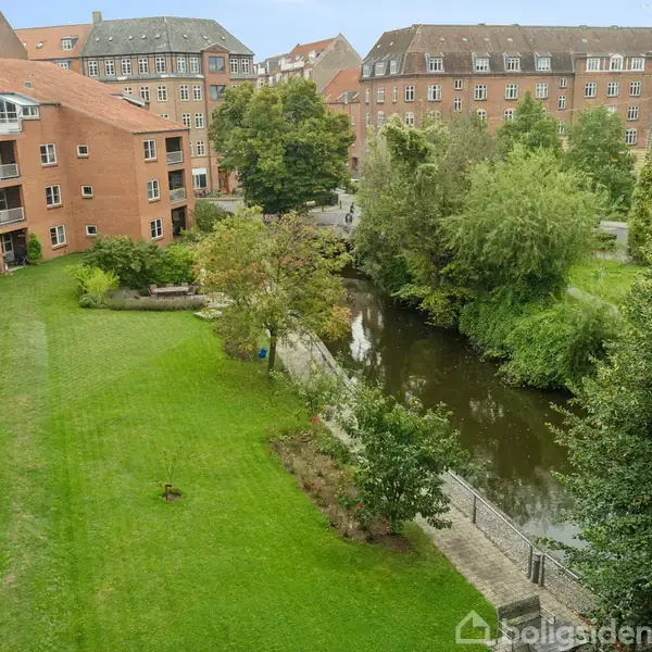 En græsplæne med træer grænser op til en flod. Bagved ses murstensbygninger med flere etager. Træer og buske omkranser området. Stemningen er stille og bynær.